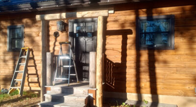 Hand-peeled log beam installed between two pine support posts above the back door, marking a major milestone in building the back porch awning at Woodsy Acres.